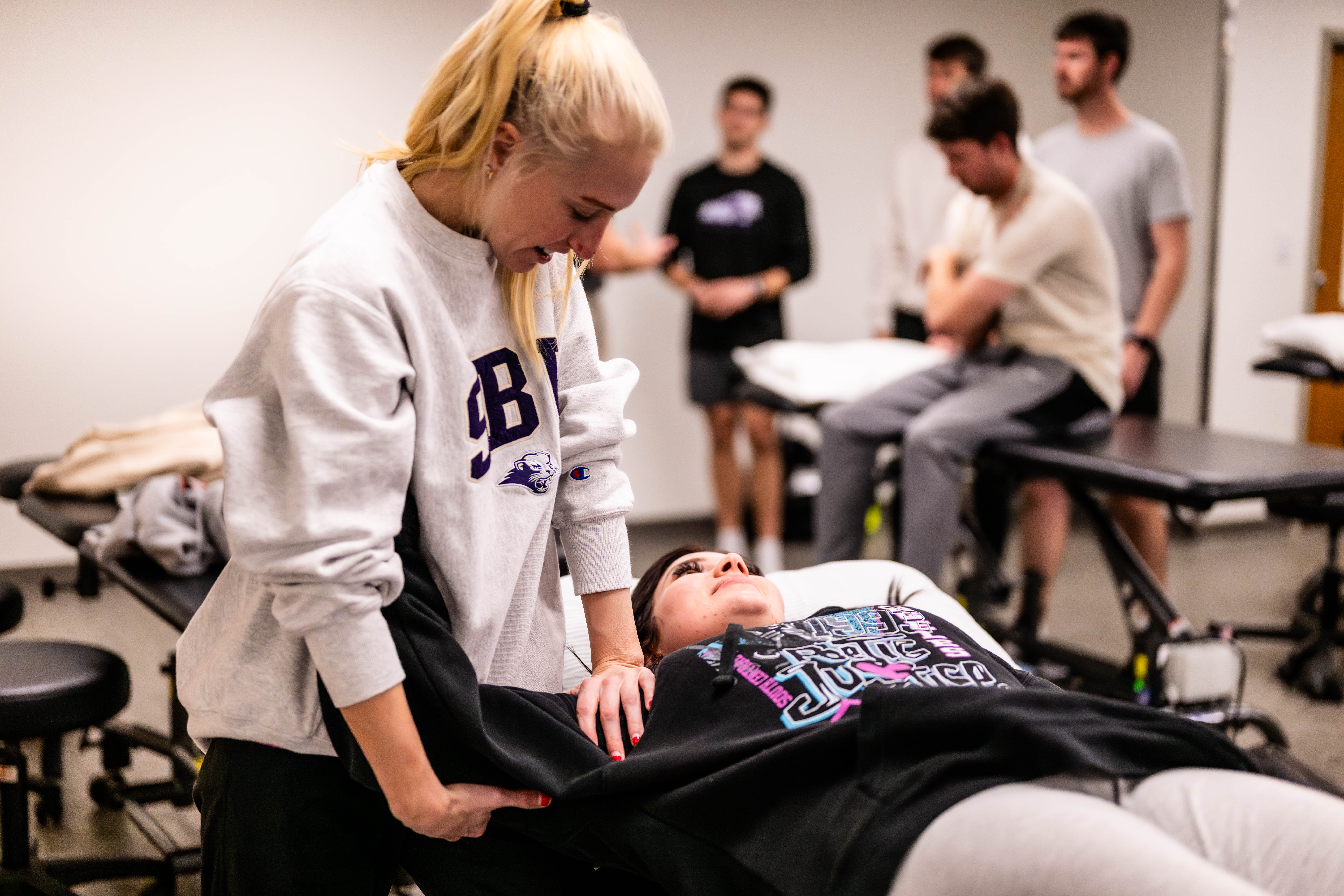 Physical therapy professor demonstrates an exercise on a student laying on lab table while explaining to another student looking on
